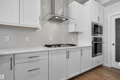 Modern kitchen featuring white shaker cabinetry, light-toned countertops, a herringbone tile backsplash, and stainless steel appliances including a built-in microwave and oven - 9432 229 Street, Edmonton, AB 