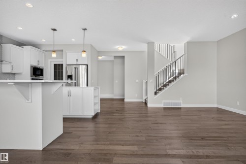 Open-concept kitchen and living area featuring wood-finish flooring, white cabinetry, stainless steel appliances, a kitchen island with pendant lighting, and a staircase with metal railings - 9432 229 Street, Edmonton, AB 