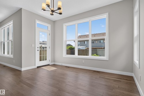Spacious room featuring wood-finish flooring, a contemporary chandelier, multiple large windows with white trim, and a glass-paneled door - 9432 229 Street, Edmonton, AB 