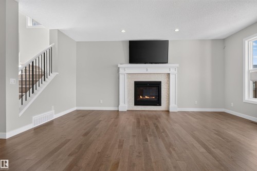 Living area featuring wood-finish flooring, a white mantel fireplace with tile surround, a mounted television, a window with white trim, and a staircase with dark balusters - 9432 229 Street, Edmonton, AB 