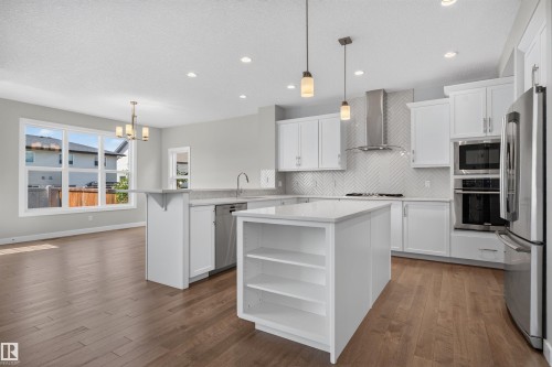 Open-concept kitchen featuring white cabinetry, stainless steel appliances, a central island with shelving, pendant lighting, and wood-finish flooring - 9432 229 Street, Edmonton, AB 
