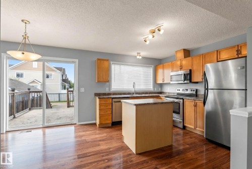 Open-concept kitchen featuring wood-finish flooring, light wood cabinetry, stainless steel appliances, and a central island with a stone-finish countertop - 7118 19A, Edmonton, AB - Indoor Photo Showing Kitchen