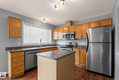 Kitchen featuring wood-finish cabinetry, stainless steel appliances, an island with countertop space, wood-finish flooring, and a large window with blinds - 7118 19A, Edmonton, AB - Indoor Photo Showing Kitchen With Double Sink