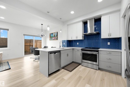 Modern kitchen featuring light wood-finish flooring, white overhead cabinetry, grey base cabinetry, stainless steel appliances, and a bold blue subway tile backsplash - 3173 Checknita Way, Edmonton, AB 