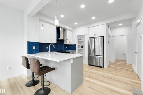 Modern kitchen featuring an island with a light-toned countertop, white cabinetry, a blue subway tile backsplash, stainless steel appliances, and light wood-finish flooring - 3173 Checknita Way, Edmonton, AB 
