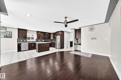 Open-concept kitchen and living area featuring rich wood-finish flooring, a contemporary ceiling fan, dark wood cabinetry, a mosaic tile backsplash, and stainless steel appliances - 16124 76 Street, Edmonton, AB 