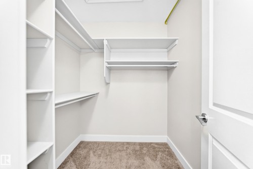 Walk-in closet featuring extensive white shelving, multiple hanging rods, neutral carpeting, and a white interior door with brushed nickel hardware - 16124 76 Street, Edmonton, AB 