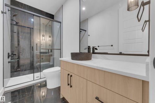 Contemporary bathroom featuring a spacious glass-enclosed shower with dark-toned tile surround, a light wood-finish vanity with a white countertop, a large framed mirror, and dark-toned fixtures throughout - 7677 Kimiwan Crescent, Edmonton, AB 