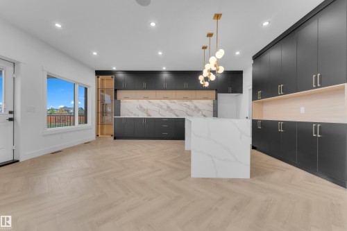 Spacious kitchen featuring two large islands with waterfall-edge countertops, extensive dark cabinetry, light wood-finish flooring in a herringbone pattern, and a modern gold-tone pendant light fixture - 7677 Kimiwan Crescent, Edmonton, AB 