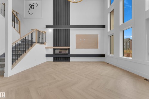 Vaulted ceiling living space featuring a modern fireplace with a black slat wall accent, oversized windows, light wood-finish herringbone flooring, and a staircase with light wood handrails and black metal spindles - 7677 Kimiwan Crescent, Edmonton, AB 