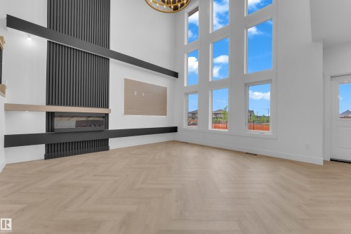 Double-height living space featuring expansive multi-pane windows, a modern linear fireplace with a textured black surround, a light wood-finish mantel, and herringbone pattern wood-finish flooring - 7677 Kimiwan Crescent, Edmonton, AB 