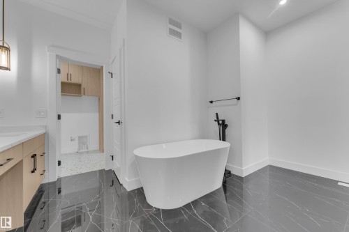 Freestanding soaking tub with a floor-mounted matte black faucet, dark stone-look flooring, and a light wood-finish vanity with a white countertop - 7677 Kimiwan Crescent, Edmonton, AB 