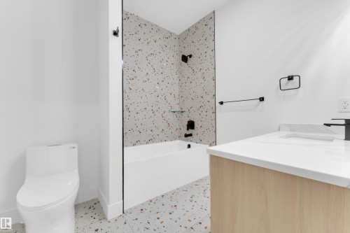 Bathroom featuring a white bathtub, speckled tile flooring and shower surround, black plumbing fixtures, and a light wood-finish vanity with a white countertop - 7677 Kimiwan Crescent, Edmonton, AB 