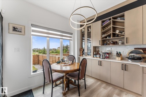 Kitchen dining area featuring a large window, light wood-finish flooring, a contemporary circular pendant light, flat-panel cabinetry with black hardware, and a tile backsplash - 94 Elm Street, Fort Saskatchewan, AB 
