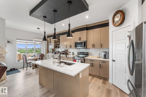 Contemporary kitchen featuring a central island with an undermount sink, light wood-finish flooring, stainless steel appliances, a white tile backsplash, and modern cabinetry with integrated wine storage - 94 Elm Street, Fort Saskatchewan, AB 