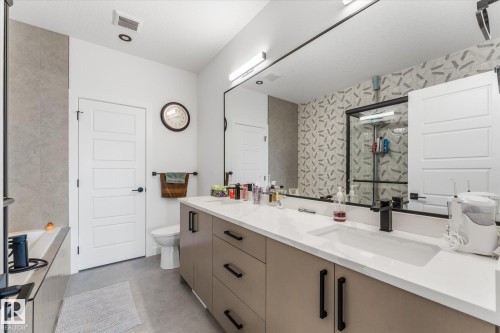 Contemporary bath featuring a dual sink vanity with a white countertop and matte black fixtures, a full-width mirror, and a tiled accent wall - 94 Elm Street, Fort Saskatchewan, AB 