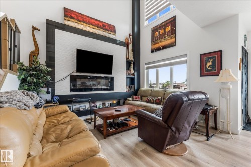 Living area featuring high ceilings, wood-finish flooring, a contemporary built-in electric fireplace, a custom accent wall with textured white panels, and multiple windows allowing abundant natural light - 94 Elm Street, Fort Saskatchewan, AB 
