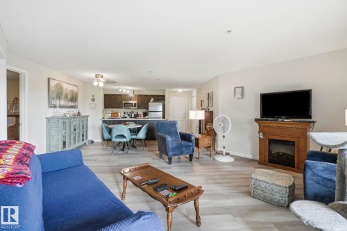 Open-concept living area featuring wood-finish flooring, a built-in electric fireplace with a wooden mantel, and a kitchen with dark cabinetry and stainless steel appliances - 1223 9363 Simpson Dr, Edmonton, AB 