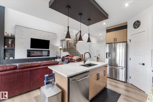 Open-concept living space featuring a kitchen island with integrated sink, stainless steel appliances, wood-finish flooring, a contemporary fireplace, and a staircase with wood handrails - 92 Elm Street, Fort Saskatchewan, AB 