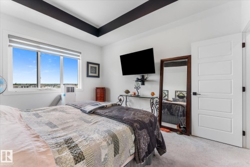 Contemporary bedroom featuring a large window, tray ceiling with dark accents, light gray carpeting, and a modern paneled door with dark hardware - 92 Elm Street, Fort Saskatchewan, AB 