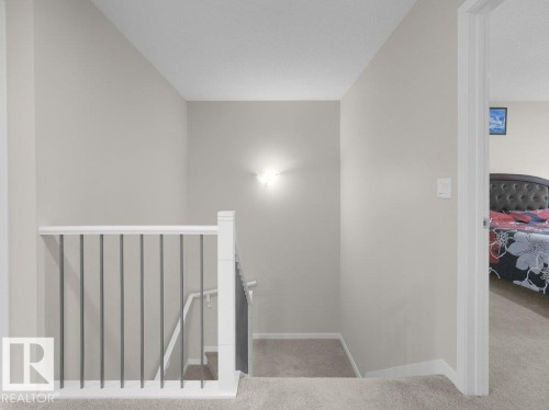 Carpeted hallway featuring a white railing with dark vertical balusters, light neutral wall paint, and a wall-mounted light fixture - 2039 13 Avenue, Edmonton, AB 