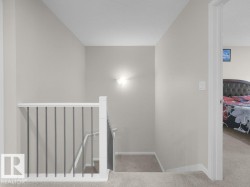 Carpeted hallway featuring a white railing with dark vertical balusters, light neutral wall paint, and a wall-mounted light fixture - 