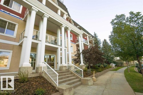 Multi-story building featuring classical columns, glass-paneled balconies, a concrete sidewalk, and mature landscaping with evergreen trees - #117 226 Macewan Sw, Edmonton, AB - Outdoor