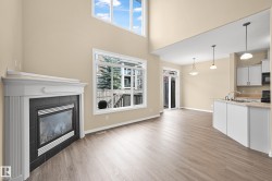 Open-concept living area featuring a fireplace with a white mantel, wood-finish flooring, high ceilings, large windows, and a kitchen with white cabinetry - 