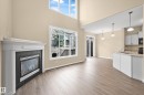 Open-concept living area featuring a fireplace with a white mantel, wood-finish flooring, high ceilings, large windows, and a kitchen with white cabinetry - 11544 11 Avenue, Edmonton, AB 