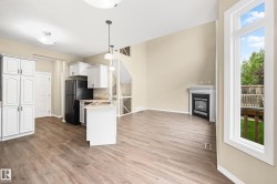 Open-concept living space featuring wood-finish flooring, a kitchen with white cabinetry and a central island, a built-in fireplace with mantel, a large window, and a visible deck area - 