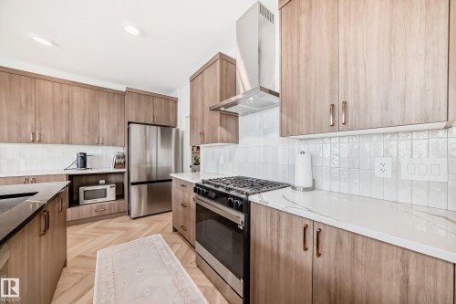 Contemporary kitchen featuring wood-finish cabinetry, stainless steel appliances, a white tile backsplash, light-toned countertops, and recessed lighting - 4231 206 Street, Edmonton, AB 