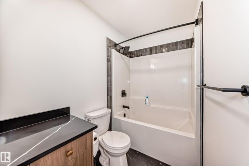 Bathroom featuring a combination bathtub and shower with dark tile surround, a toilet, a vanity with a dark countertop and light-colored cabinetry, and dark flooring - 4231 206 Street, Edmonton, AB 