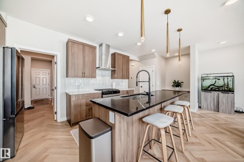 Modern kitchen featuring a large island with a dark countertop and integrated sink, herringbone wood-finish flooring, wood-grain cabinetry, a white subway tile backsplash, and stainless steel appliances including a range hood - 4231 206 Street, Edmonton, AB 