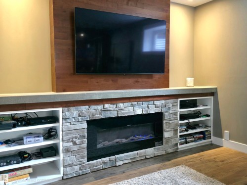 Built-in entertainment wall featuring a stone-clad electric fireplace, wood-finish accent wall, integrated display shelving, and a concrete-finish mantel - 1 Hemlock Close, Fort Saskatchewan, AB 