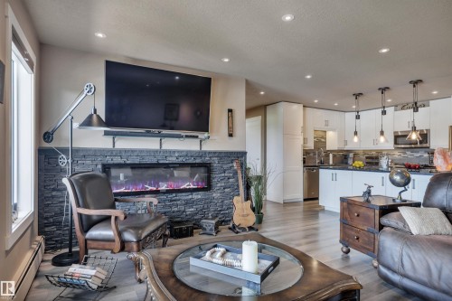 Open-concept living area featuring an electric fireplace with a stacked stone surround, recessed lighting, wood-finish flooring, white kitchen cabinetry, and stainless steel appliances - 305 5212 48 Avenue, Wetaskiwin, AB 