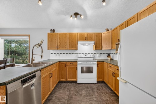 Bright kitchen featuring natural wood cabinetry, dark countertops, a stainless steel dishwasher, and a double basin sink with a modern faucet - 1 Primrose Boulevard, Sherwood Park, AB 