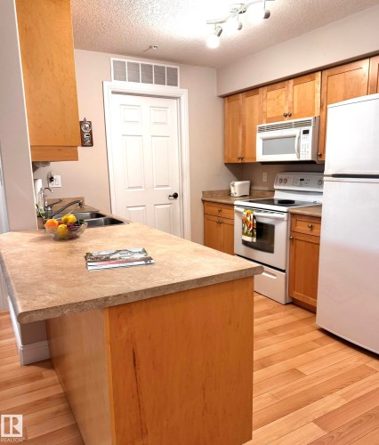 Kitchen featuring a central island with stone-finish countertop, wood-finish flooring, light wood cabinetry, track lighting, and a white appliance suite - 330 13111 140 Avenue, Edmonton, AB 