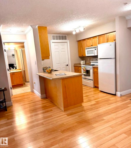Kitchen area featuring a light wood-finish island with a light-toned countertop, ample natural wood cabinetry, and a suite of white appliances - 330 13111 140 Avenue, Edmonton, AB 