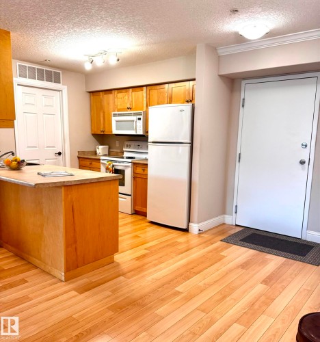 Kitchen area featuring wood-finish flooring, light wood cabinetry, white appliances, and a central island with a sink - 330 13111 140 Avenue, Edmonton, AB 