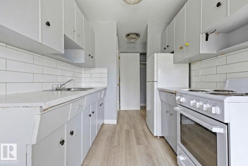 Galley kitchen featuring white cabinetry, subway tile backsplash, light-toned wood-finish flooring, white appliances, and a stainless steel sink - 15 10164 150 Street, Edmonton, AB 