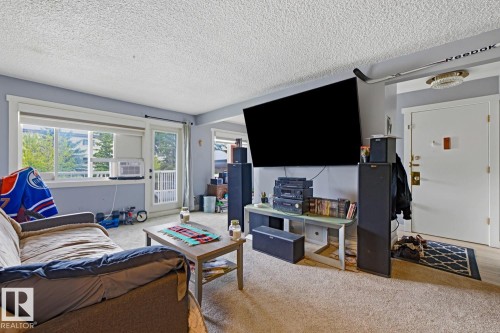 Carpeted living area featuring a neutral wall color, textured ceiling, and a large window with sliding glass door - 15 10164 150 Street, Edmonton, AB 