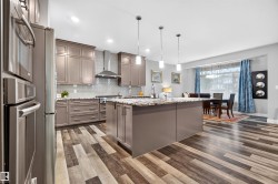 Open-concept kitchen featuring wood-finish flooring, a large island with pendant lighting, stainless steel appliances, taupe cabinetry, and granite countertops - 