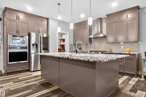 Spacious kitchen featuring a large island with a stone countertop, extensive cabinetry, stainless steel appliances, a tile backsplash, and wood-finish flooring - 1603 158 Street, Edmonton, AB 