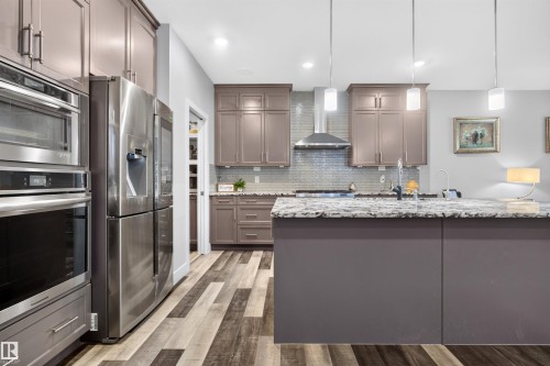 Contemporary kitchen featuring a large island with a speckled countertop, stainless steel appliances, modern pendant lighting, shaker-style cabinetry, and wood-finish flooring - 1603 158 Street, Edmonton, AB 
