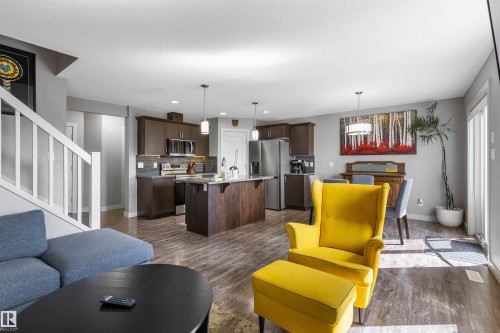 Spacious open-concept living area featuring wood-finish flooring, a kitchen with dark cabinetry and stainless steel appliances, a central island with pendant lighting, a dining area with a modern chandelier, and a white staircase with a railing - 1651 Hammond Crescent, Edmonton, AB 