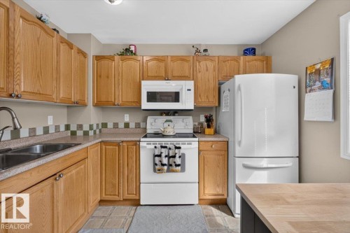 Functional kitchen featuring wood cabinetry, white appliances, dual basin sink, and a tiled backsplash - 27 Walden Park, St. Albert, AB 