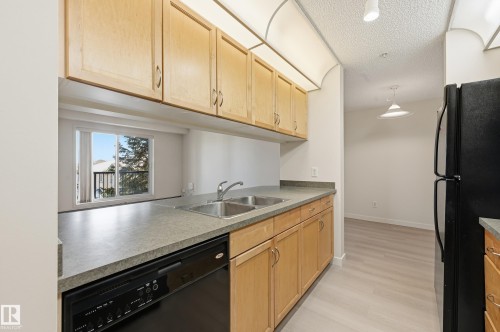 Kitchen featuring light wood-finish cabinetry, black appliances, a double basin sink, and a countertop extending to a pass-through window - 314 2430 Guardian Road, Edmonton, AB 