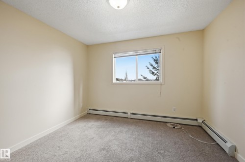 Neutral-toned room featuring a single window with white trim, light beige wall paint, gray carpeting, and baseboard heating units - 314 2430 Guardian Road, Edmonton, AB 