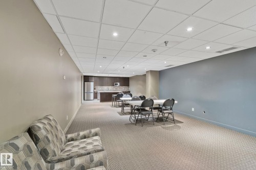Spacious communal area with patterned carpeting, recessed lighting, drop ceiling, and a built-in kitchenette featuring dark wood cabinetry and a stainless steel refrigerator - 201 10388 105 Street, Edmonton, AB 