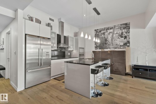 Modern kitchen featuring stainless steel appliances, a center island with pendant lighting, dark countertops, white cabinetry, and wood-finish flooring - 201 10388 105 Street, Edmonton, AB 